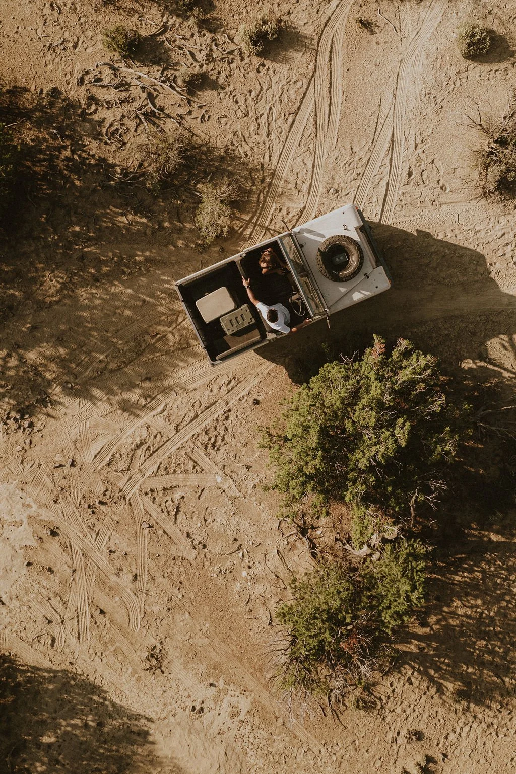 An aerial view of a desert landscape with a white off-road vehicle and two people inside. The terrain has tire tracks, bushes, and small trees.