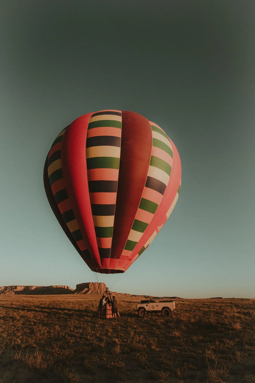 A colorful hot air balloon with striped patterns in red, green, beige, and black is about to take off in a desert landscape with a clear sky. Three people are standing near the basket, and a white vehicle is parked nearby.
