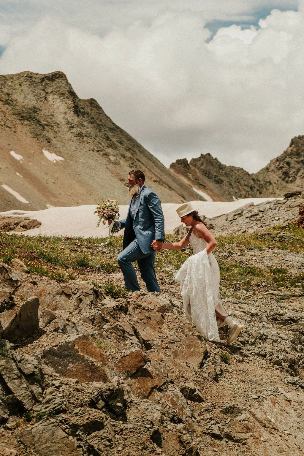 A couple dressed in wedding attire hike through a rocky mountain landscape with snow patches and cloudy sky, holding hands. The groom wears a blue suit and holds a bouquet, while the bride wears a white dress and a wide-brimmed hat.