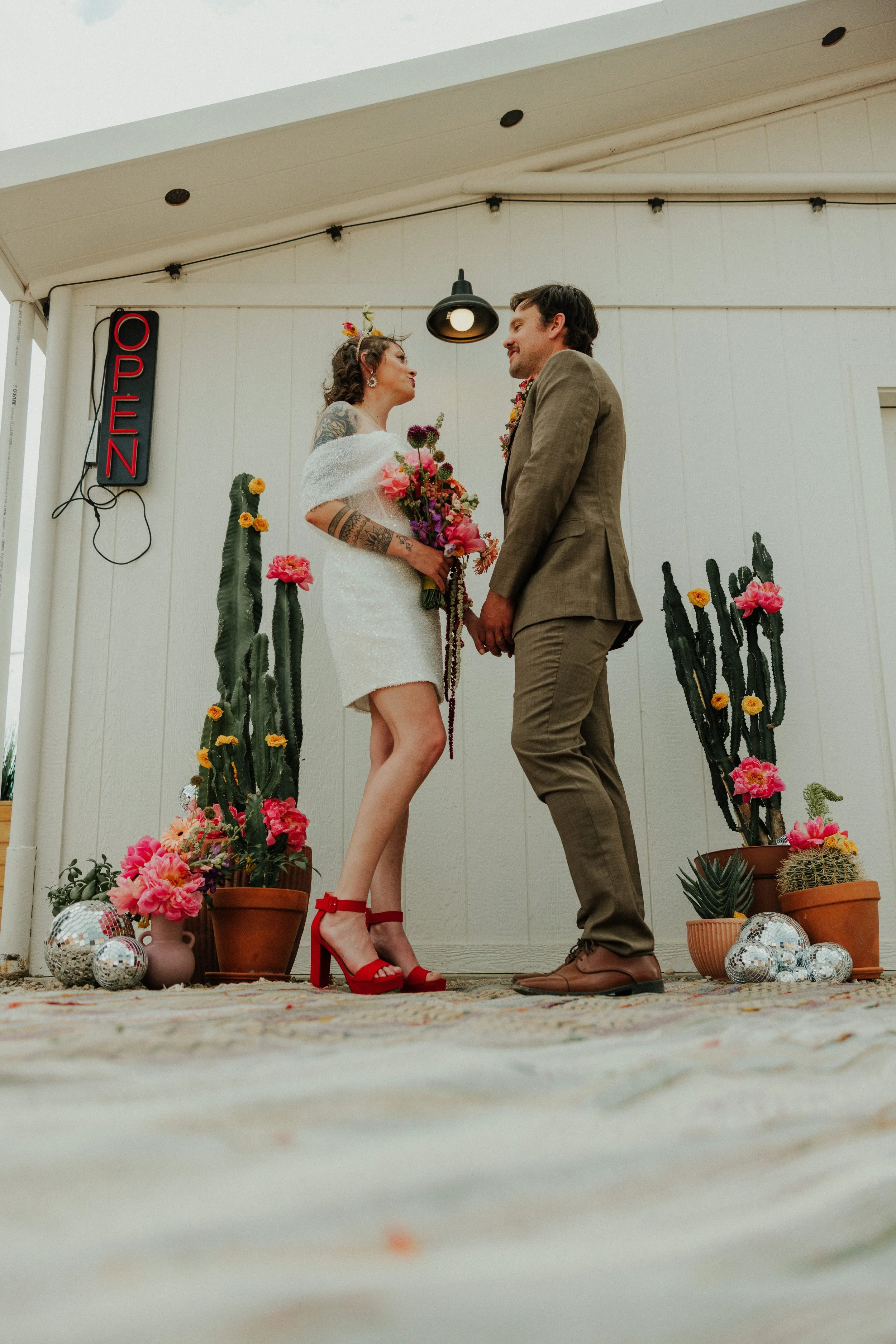 A bride and groom holding hands during their wedding ceremony, standing on an outdoor decorated area with cacti and pink flowers, with an open sign nearby.