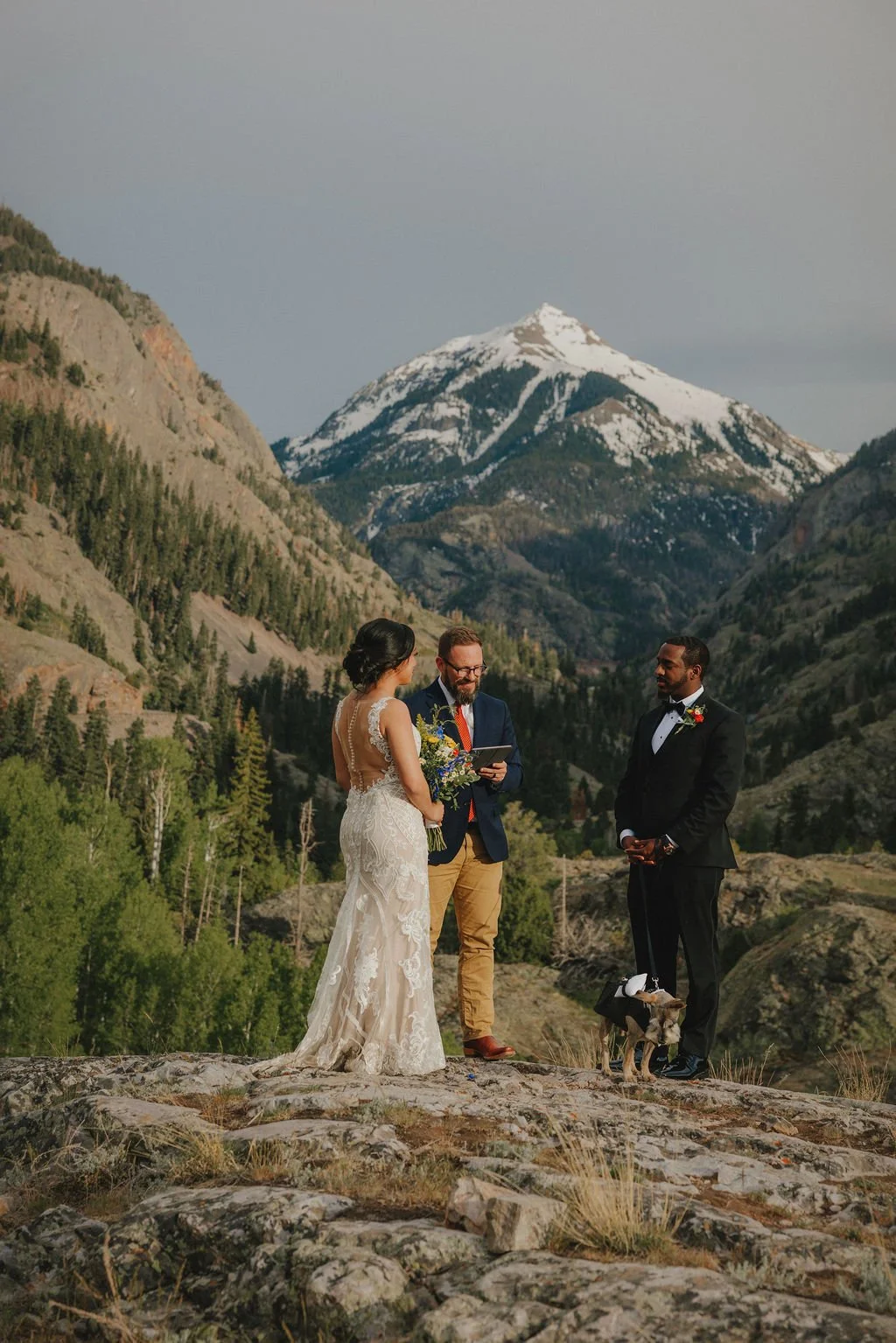 A wedding ceremony is taking place outdoors on a rocky terrain with a mountain and snowy peak in the background. A woman in a white wedding dress, a man in a black tuxedo, and another man in a blue blazer and khakis are participating, with the woman 