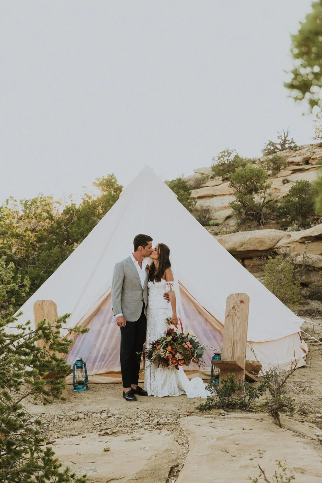A couple in wedding attire kissing in front of a white tent in a desert-like landscape with rocks and sparse trees, with lanterns and a bouquet of flowers at the bride's side.