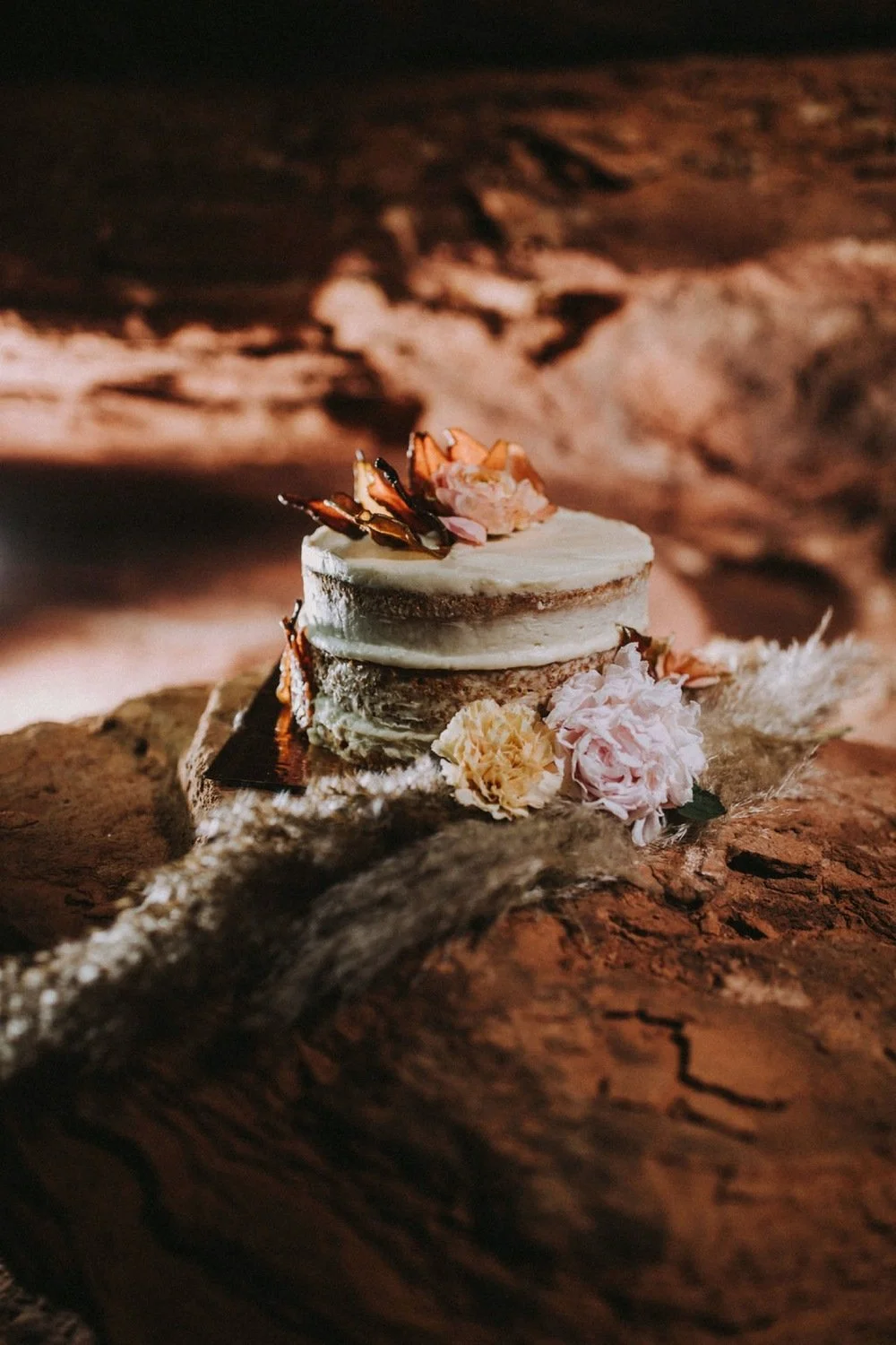 A small, layered cake with white frosting, decorated with dried flowers and petals, placed on a rustic wooden surface against a dark, cloudy sky background.