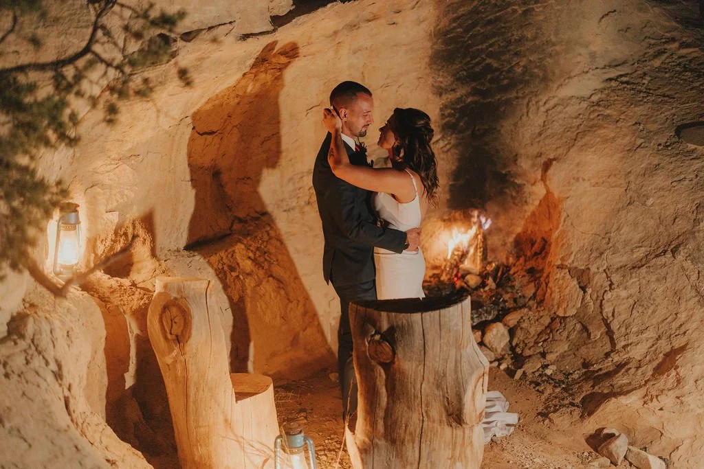 A couple in wedding attire stands close and embraces inside a rustic cave, with a small fire on a piece of wood in front of them and warm lighting creating a romantic atmosphere.