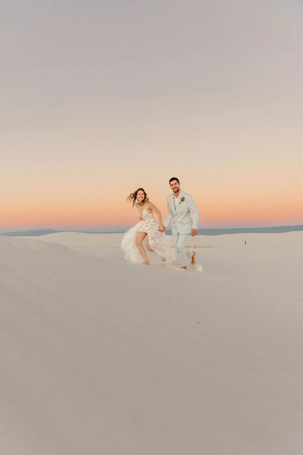 A bride and groom in wedding attire holding hands and smiling while walking in a sandy desert at sunset.