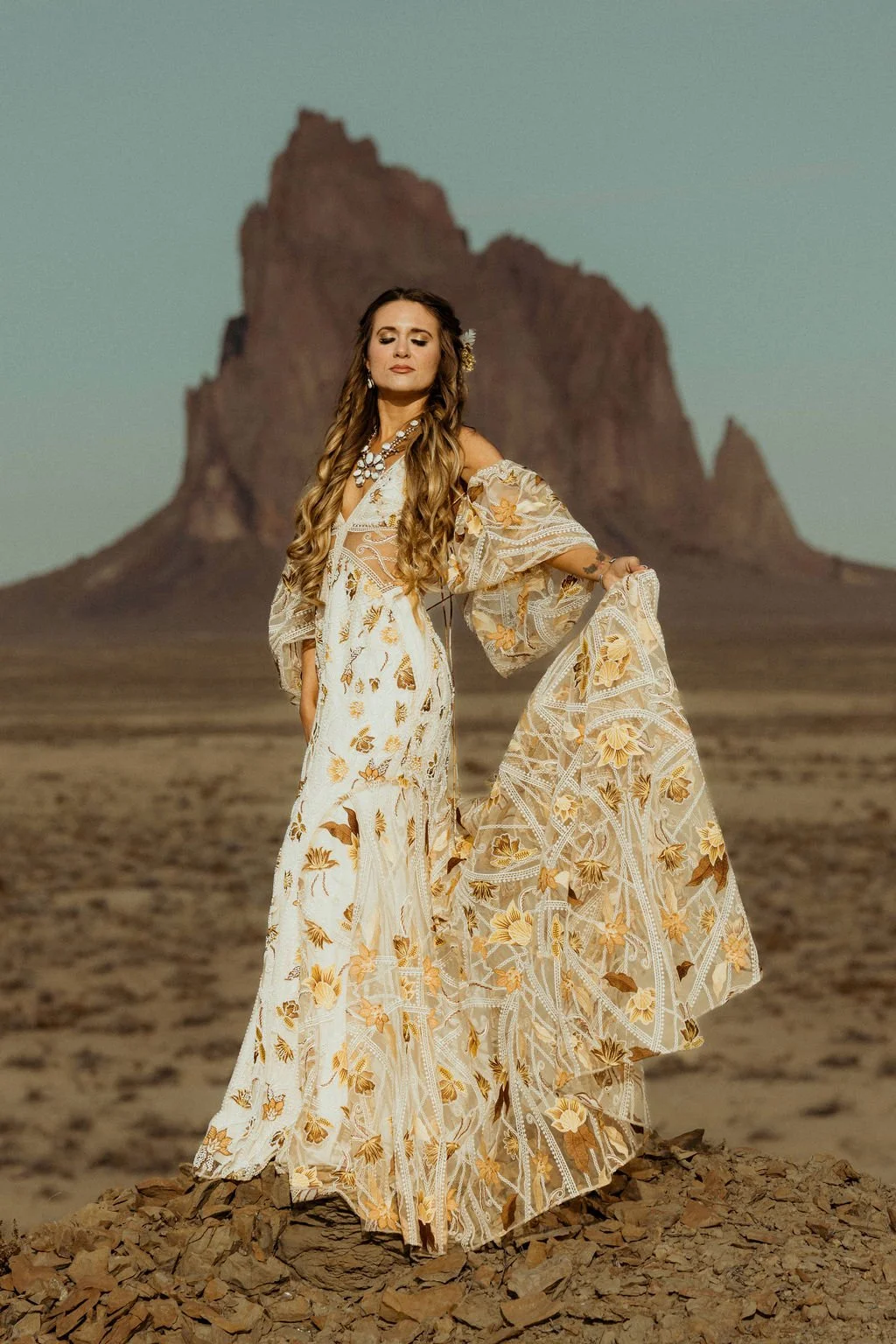 A woman in an elegant, floral dress with flowing sleeves stands on rocky terrain in front of a desert landscape featuring large, rugged mountain formations in New Mexico.