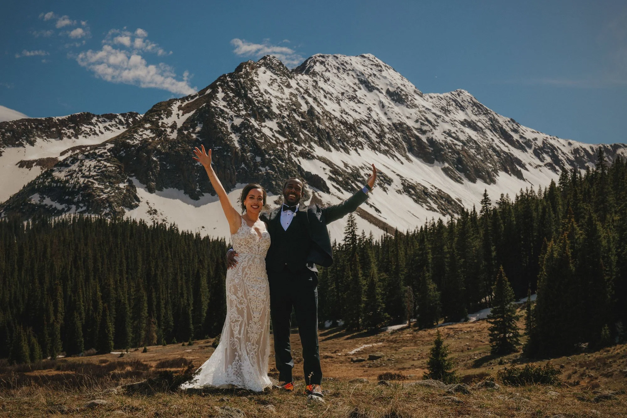 Wedding couple standing outdoors in front of a mountain range with snow-capped peaks and evergreen trees in Colorado, celebrating with arms raised.