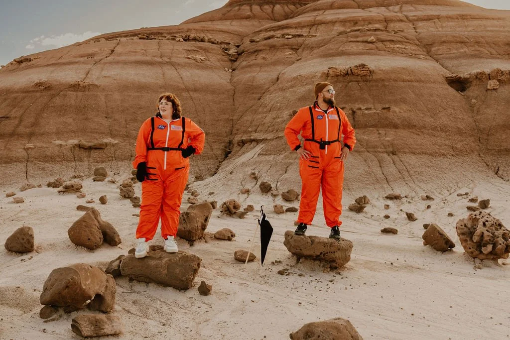 A couple dressed as astronauts in orange space suits standing on rocks in the Utah desert with a large cliff in the background.