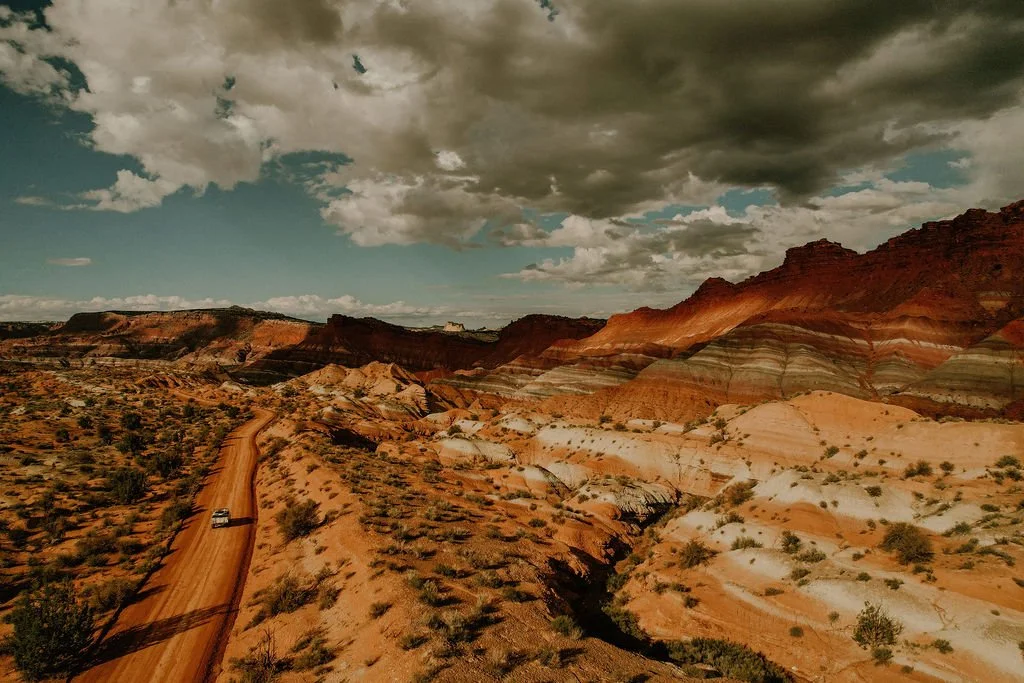 A dirt road winding through a desert landscape with multicolored layered rock formations under a partly cloudy sky.