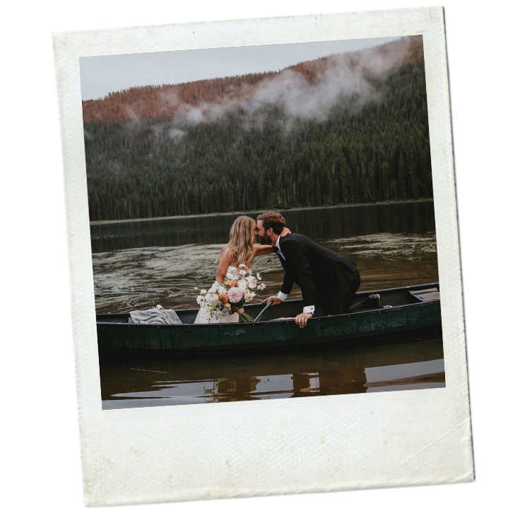 A newlywed couple in wedding attire sharing a kiss in a boat on a calm lake, with forested mountains and mist in the background.