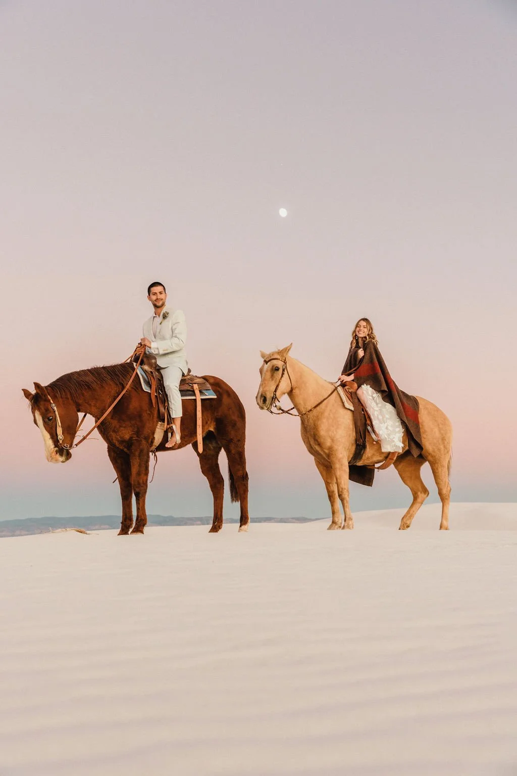 Man and woman riding horses on white sandy terrain during dusk, with the moon visible in the sky.