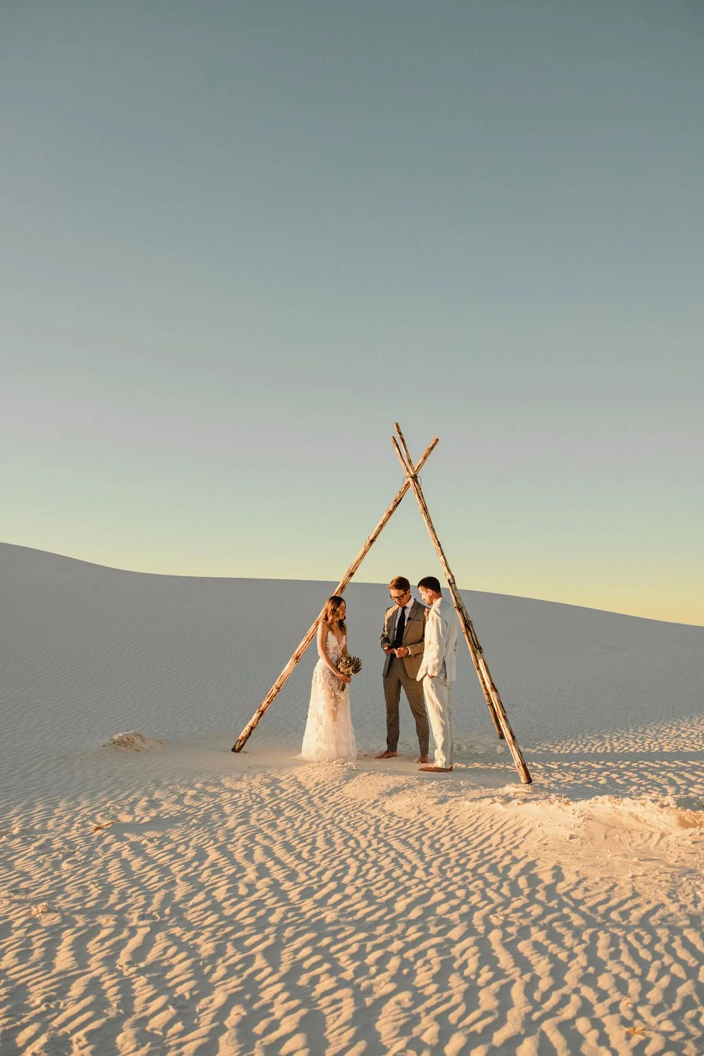A wedding ceremony taking place in a desert, with a simple wooden tent structure and a bride and groom standing under it, an officiant. The bride is wearing a white lace gown and holding a bouquet, while the groom is in a light suit. The sky is clear