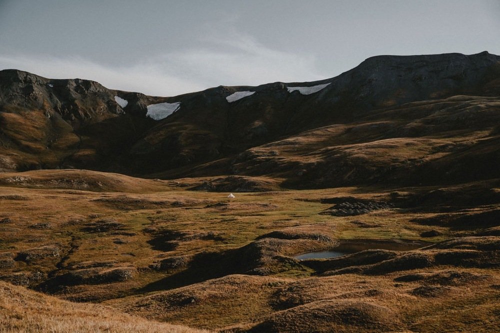A vast mountainous landscape with rolling hills, patches of snow on the mountain peaks, and a small tent in the distance, under a cloudy sky.
