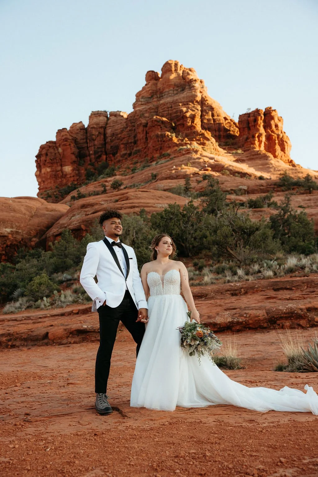 A wedding couple standing hand in hand on red desert terrain with large red rock formations in the background, the groom in a white tuxedo jacket and black pants, the bride in a white gown holding a bouquet.