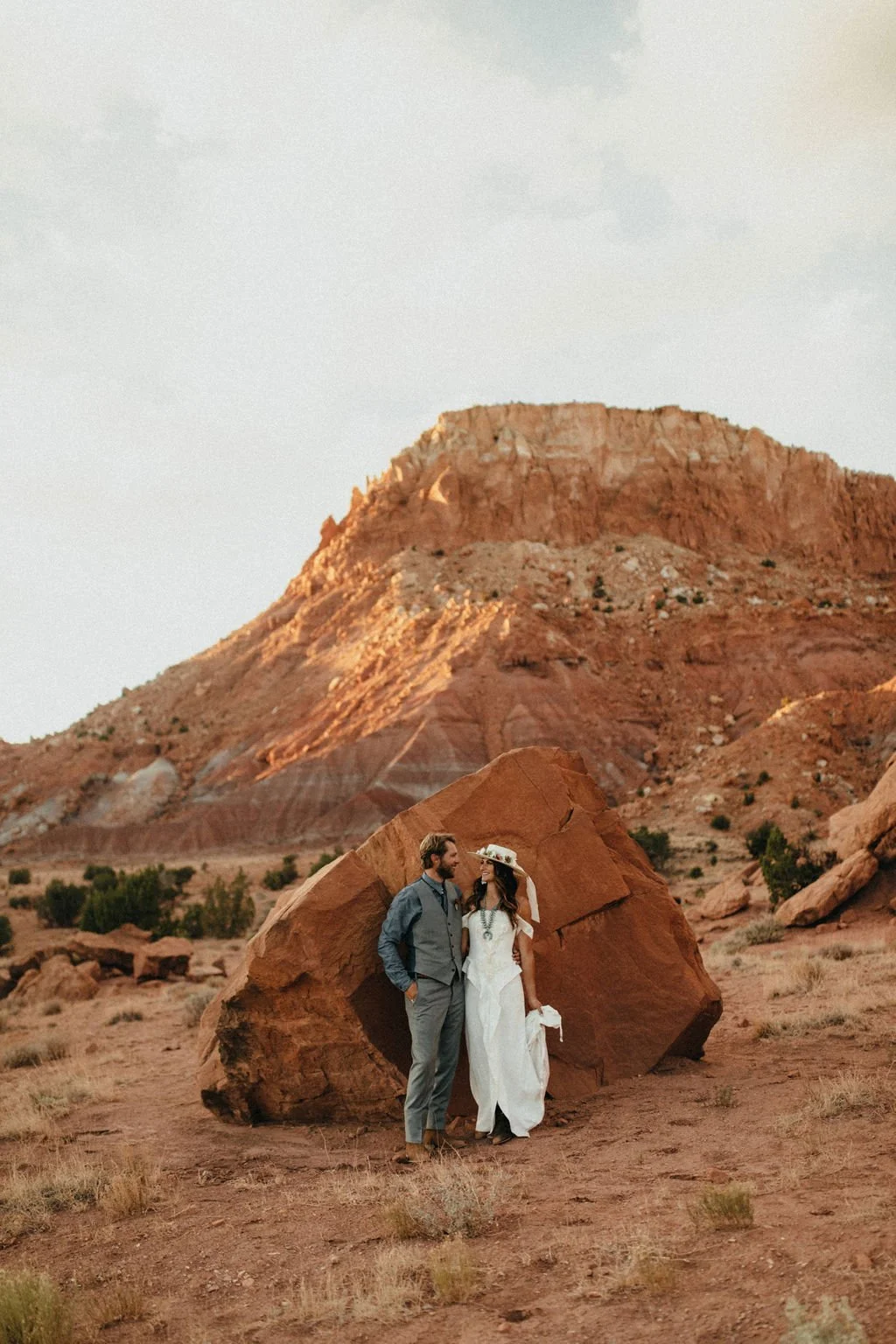 A couple stands close together in a desert landscape with reddish rock formations, one wearing a gray vest and pants and the other in a white dress with a wide-brimmed hat.