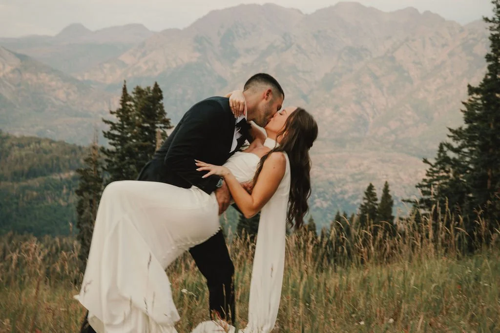 A couple in wedding attire sharing a kiss outdoors in a mountainous landscape.