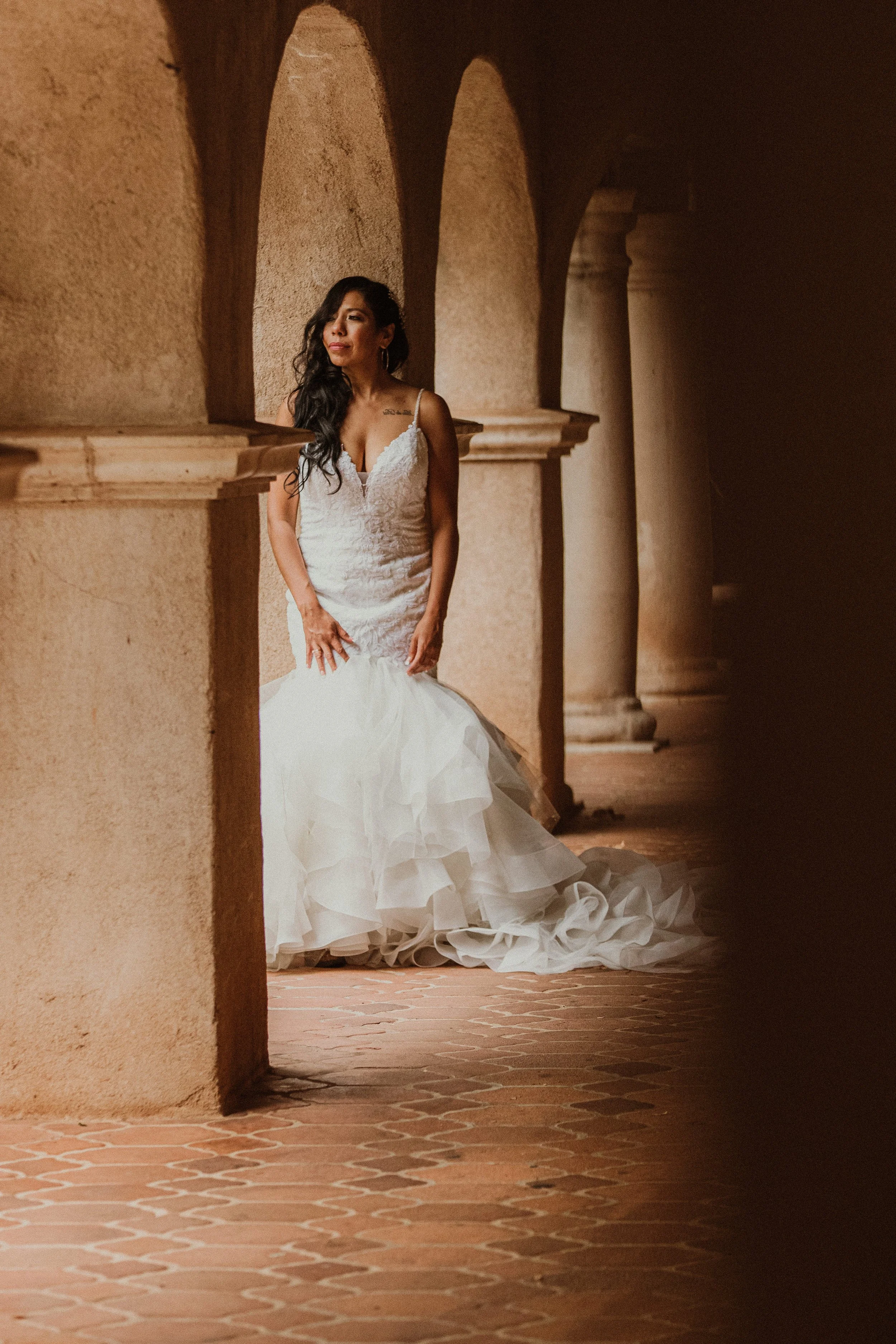 A woman in a white wedding gown standing in a hallway with stone arches and columns, with sunlight illuminating her face and dress.