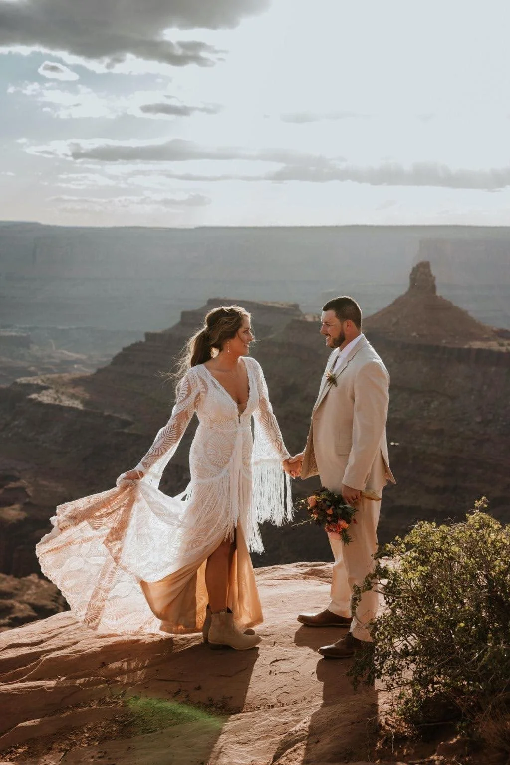 A bride and groom holding hands and smiling at each other in a desert landscape with rock formations and a cloudy sky.
