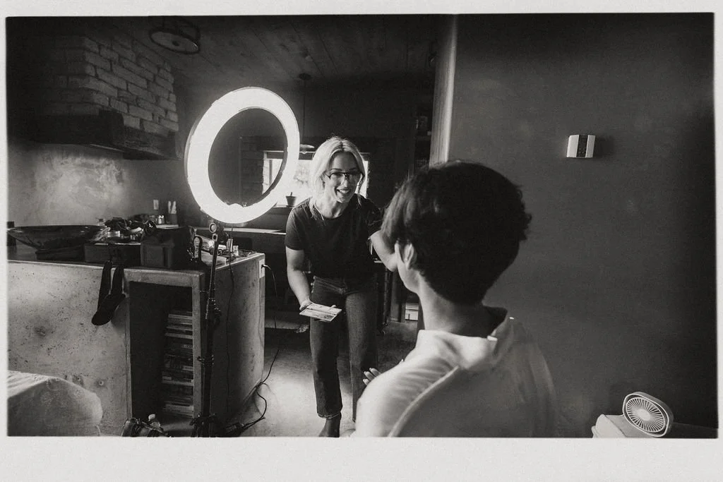 A woman with glasses and short hair smiling and holding papers, standing in a room with a large illuminated ring light, talking to a seated person with short hair.