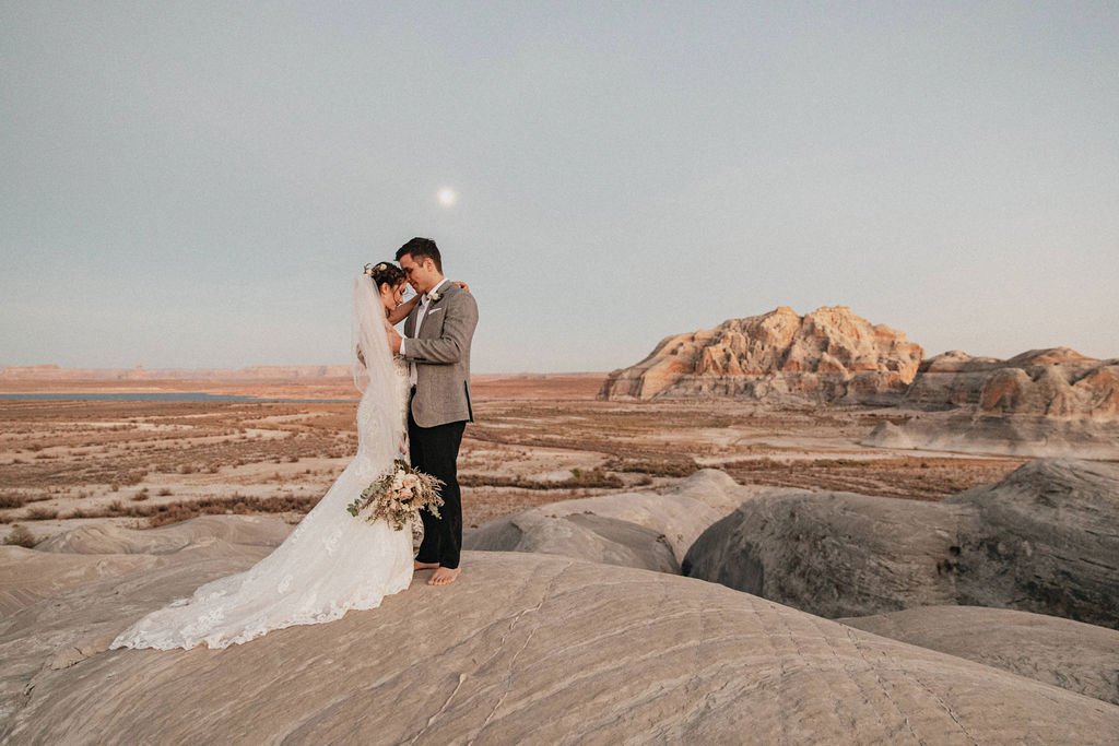 A bride and groom standing on rocks in a desert landscape at sunset, embracing, with mountains in the background and the moon visible in the sky.