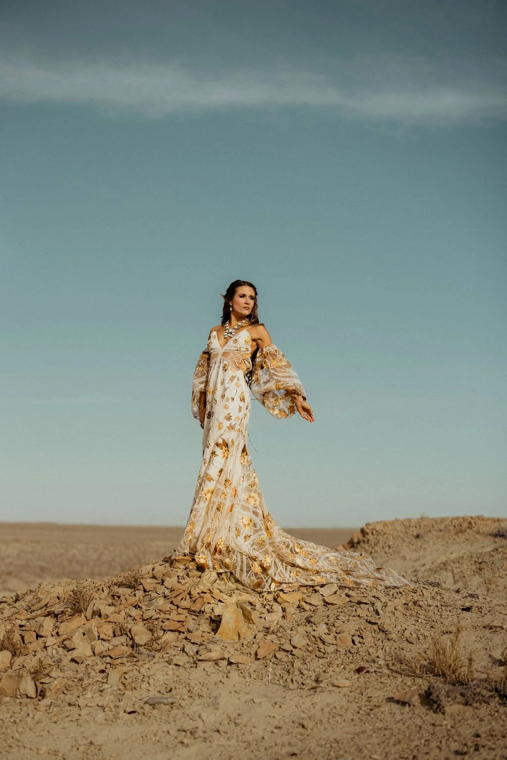 A woman standing on rocks in a desert wearing a long, flowing, patterned dress with off-shoulder sleeves and jewelry, under a partly cloudy sky.