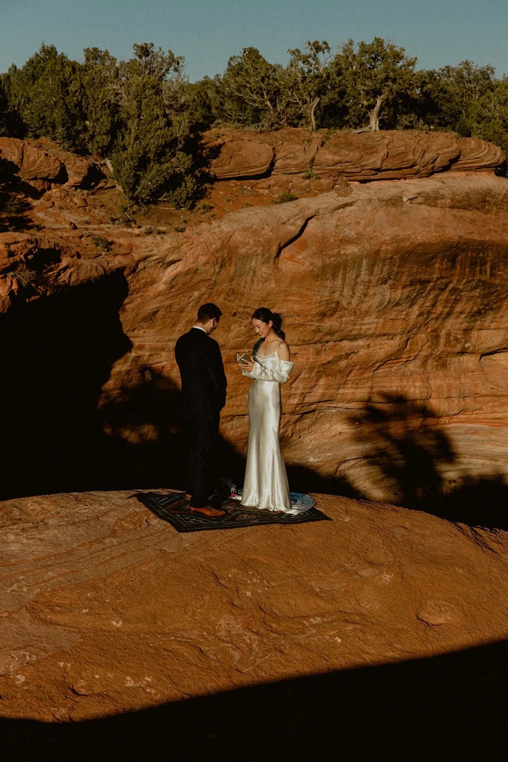A couple dressed in formal wedding attire standing on a small rug in a desert landscape with red rocks and trees in the background, during the daytime.