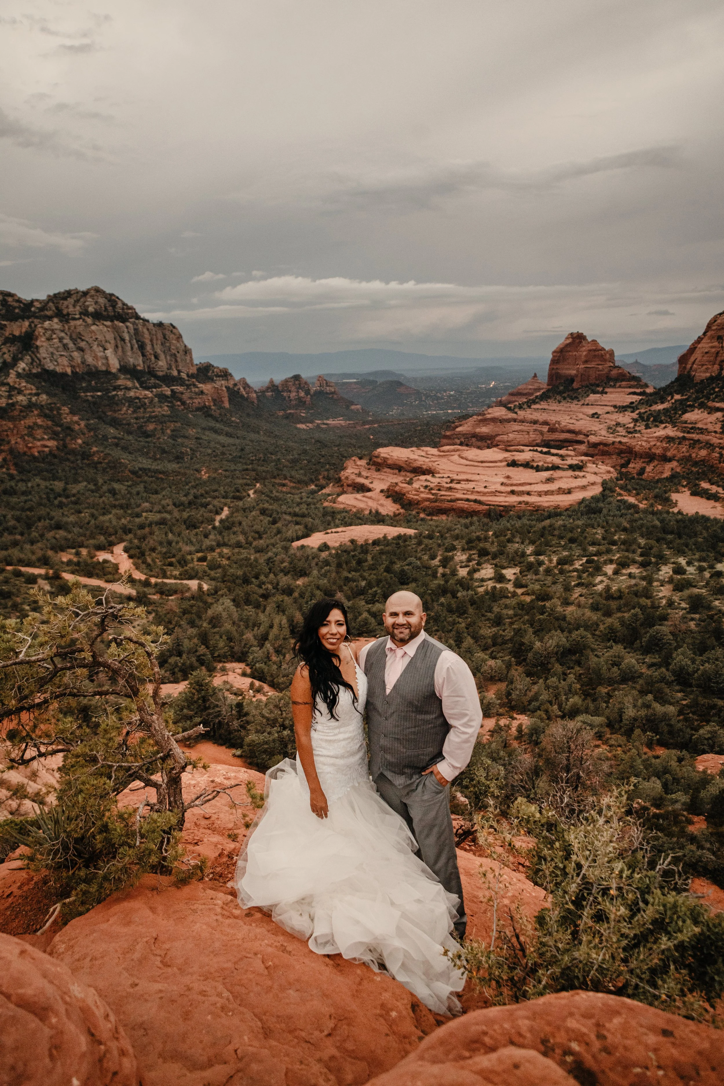 A bride and groom standing on a rocky ledge in a desert landscape with red rock formations and greenery in the background.