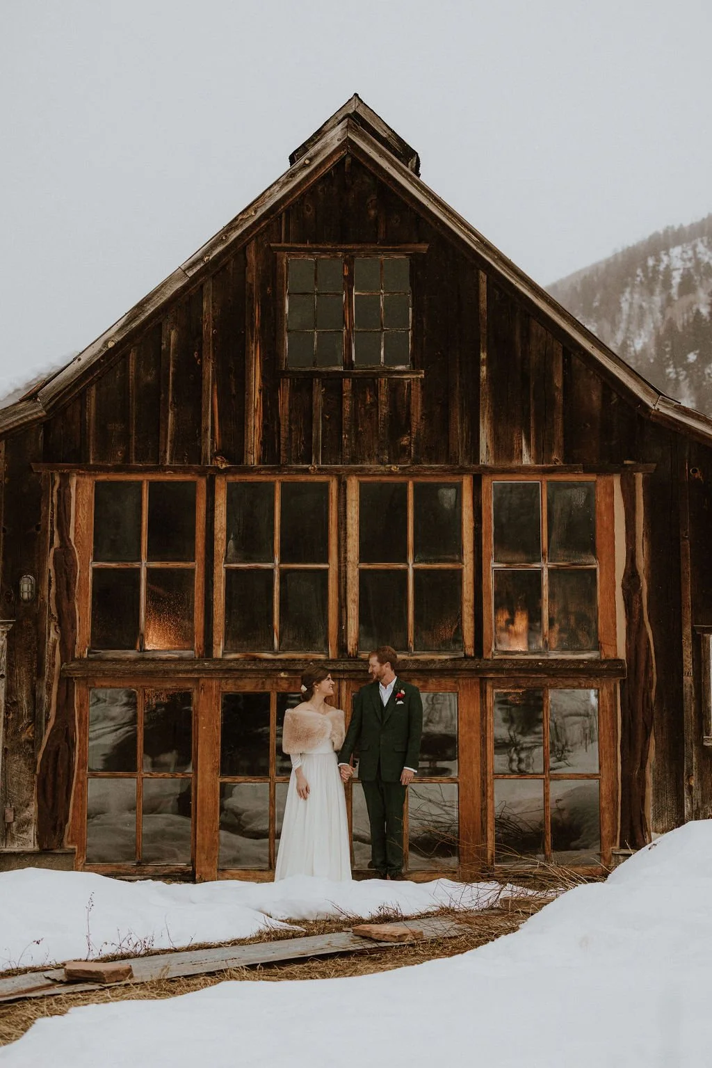 A bride and groom holding hands and looking at each other in front of a rustic wooden building with large glass windows, surrounded by snow, in a mountainous area.