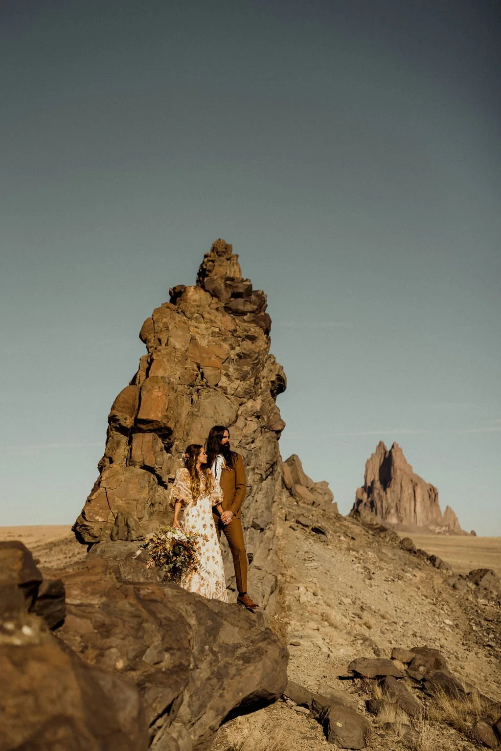 A couple standing on rocks in a desert landscape with large rock formations in the background, during late afternoon or sunset.