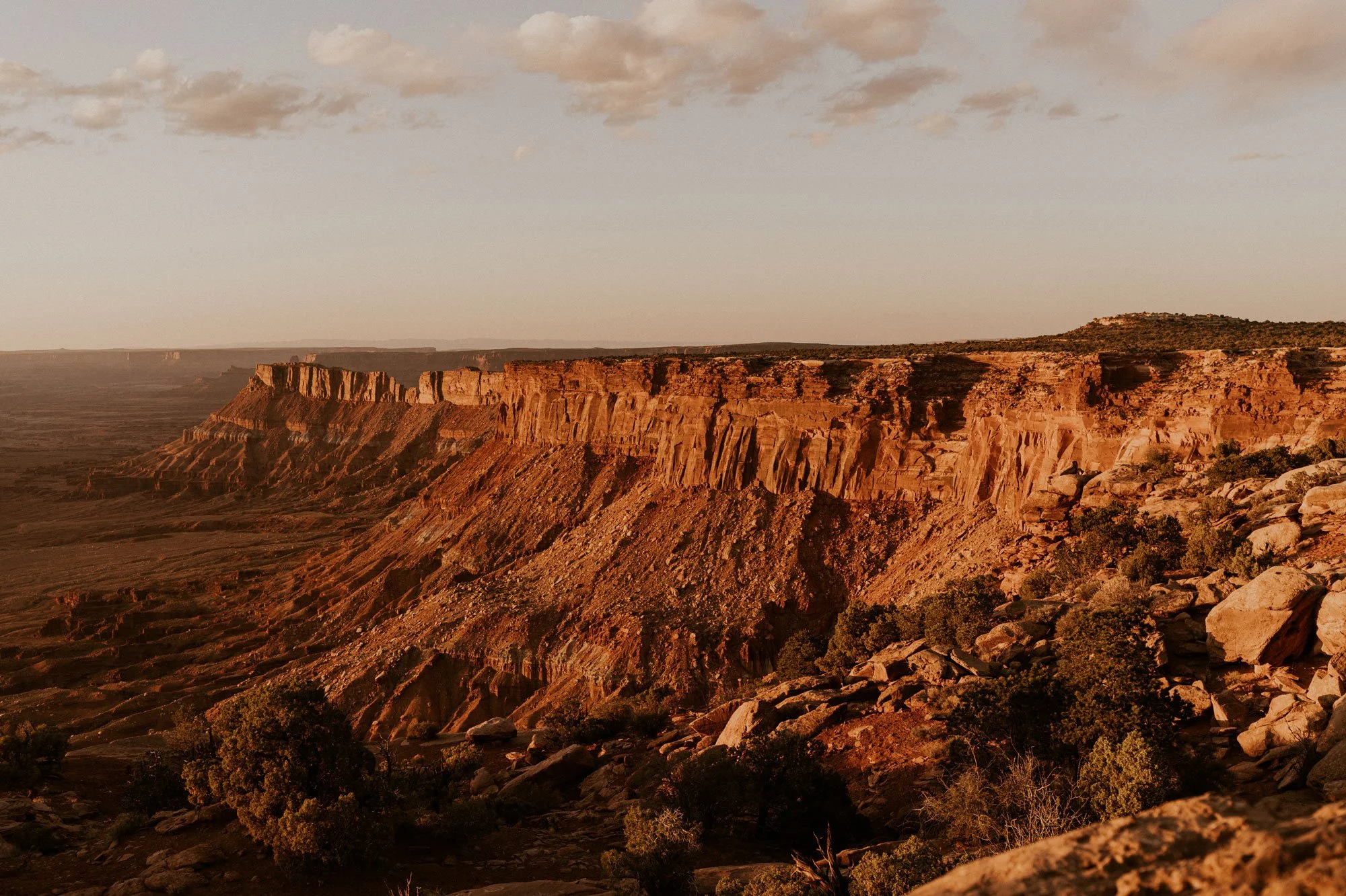Sunset over a canyon with layered rock formations and scattered shrubs in the American Southwest.