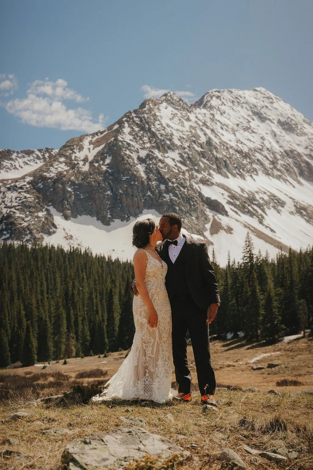 A couple dressed in wedding attire kissing outdoors with a snow-capped mountain and pine trees in the background.