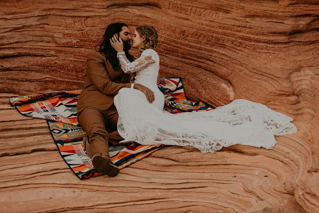 A couple sits on a colorful blanket in a natural rock formation, embracing. The woman wears a white lace dress, and the man wears a brown suit.