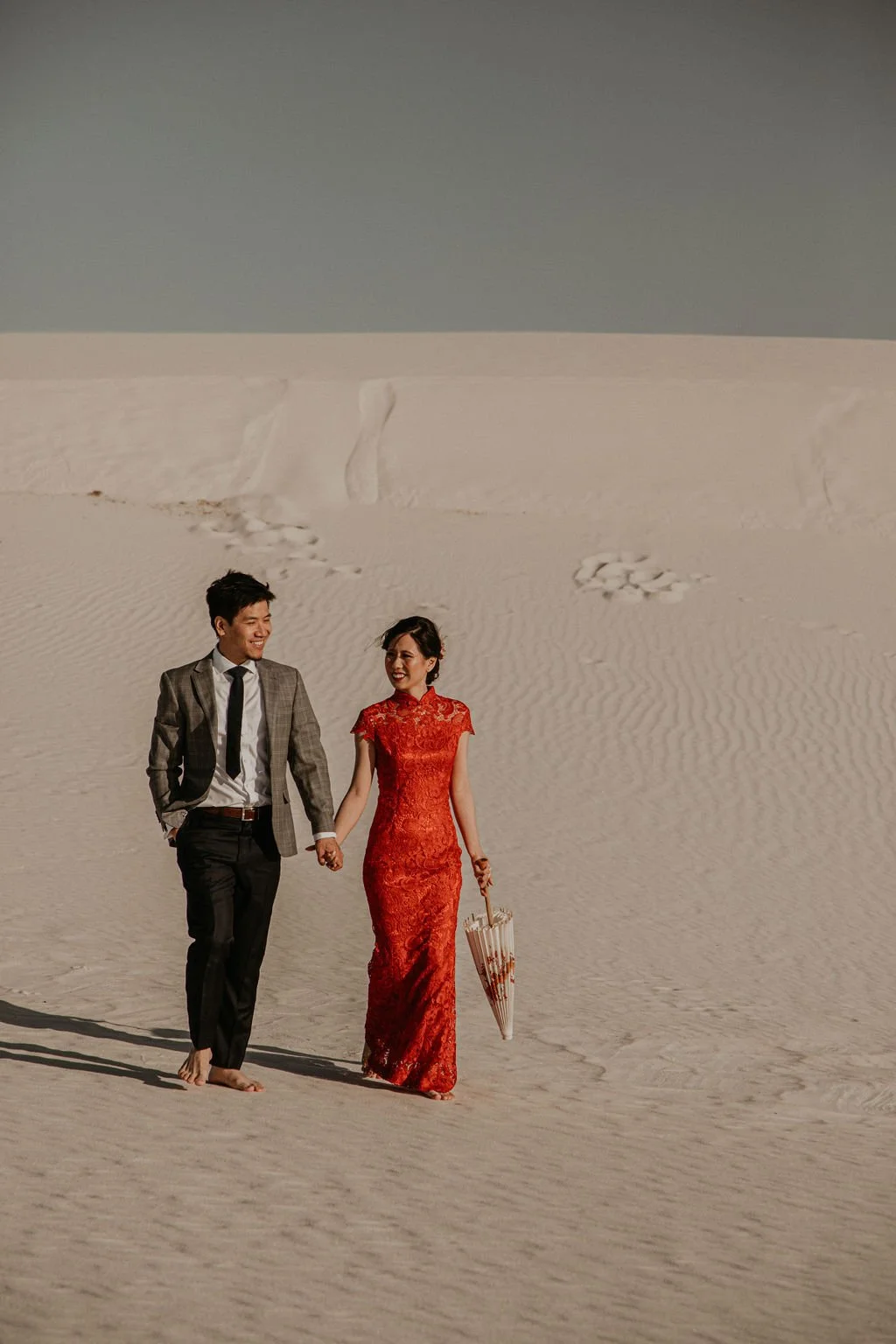 A couple walking hand in hand barefoot on a sandy desert, the woman wearing a red dress and holding an umbrella, the man in a suit.
