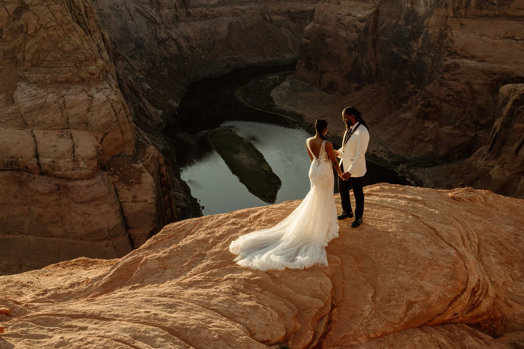 A bride and groom holding hands on a large rock formation overlooking a winding river at Horseshoe Bend at sunset.