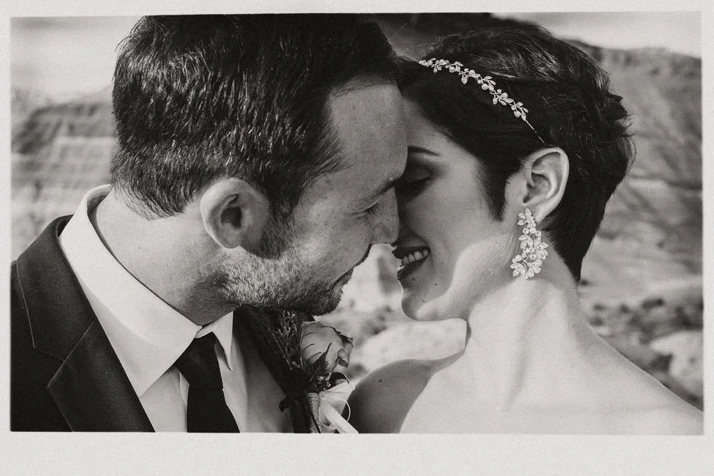 A black and white photo of a couple with their foreheads touching, smiling, and about to kiss, on what appears to be a scenic outdoor location.