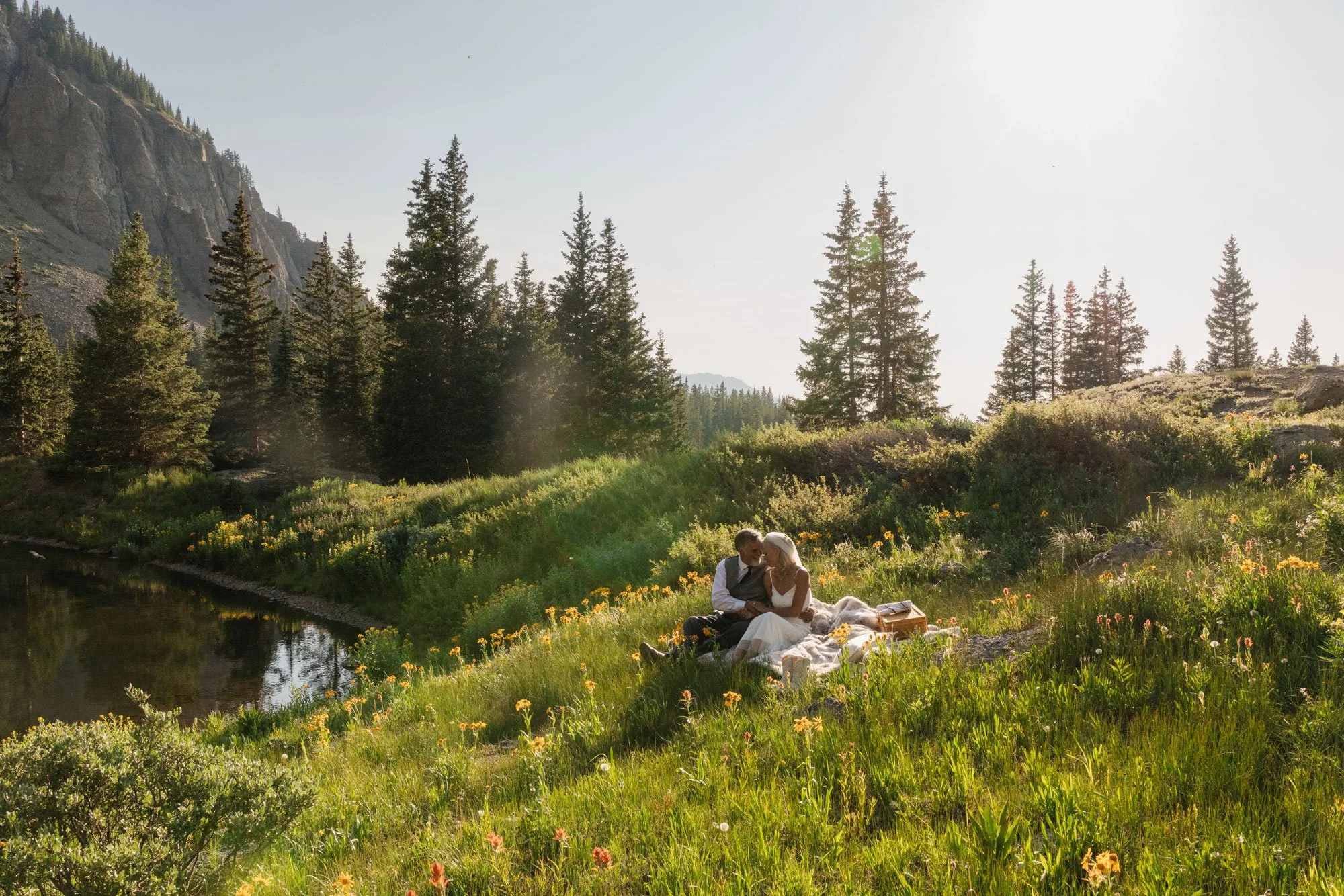 A couple sitting on a blanket in a meadow next to a small river, surrounded by wildflowers and tall trees, with mountains in the background. They are dressed in formal wedding attire.