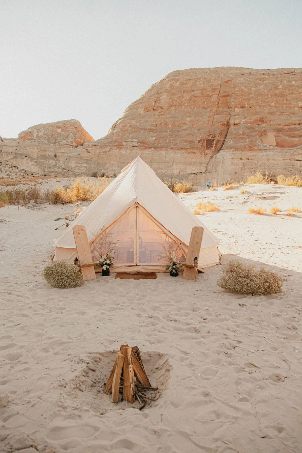 A white canvas tent set up in a sandy desert with rocky formations in the background, decorated with flowers, and a small campfire in front.