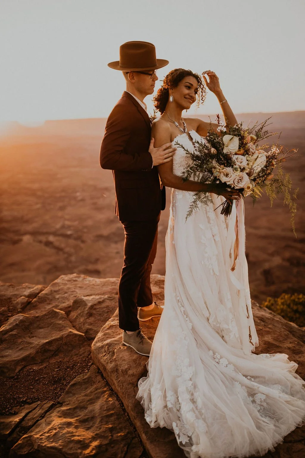 A bride and groom standing on a rocky ledge during sunset. The bride holds a bouquet of flowers and is wearing a white wedding dress, while the groom is dressed in a dark suit and a brown hat, standing close together.