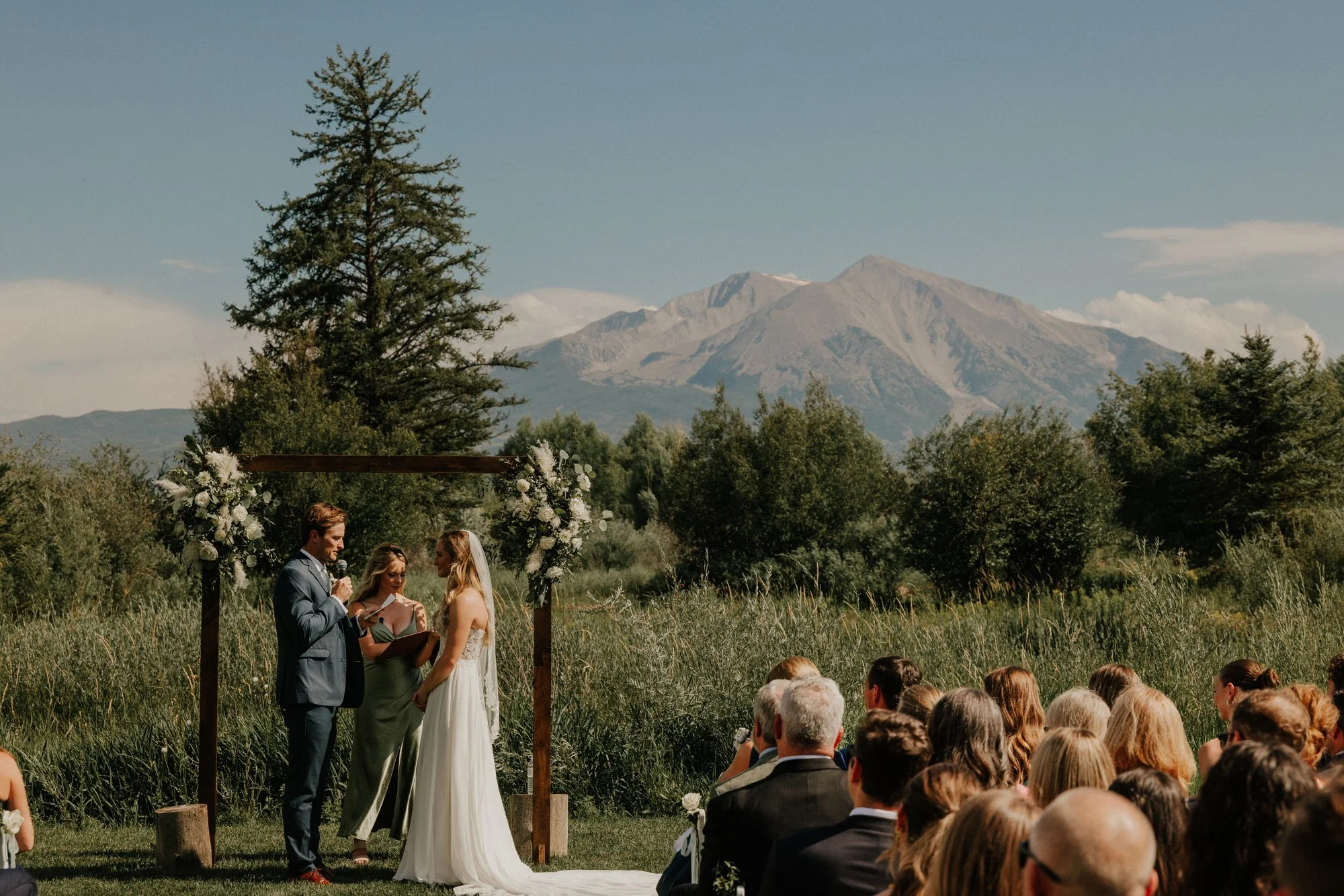 A wedding ceremony officiated outdoors with mountains in the background in Colorado, featuring a couple at an altar with floral decorations, and guests seated on the grass.
