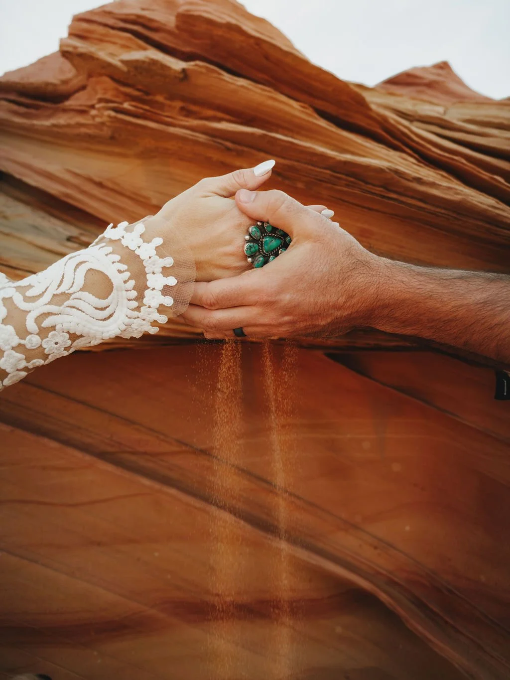 Two hands, one adorned with a large turquoise ring and the other with a white lace sleeve, hold each other over a canyon with layered red rock formations. Sand is falling from the hand with the ring into the canyon.