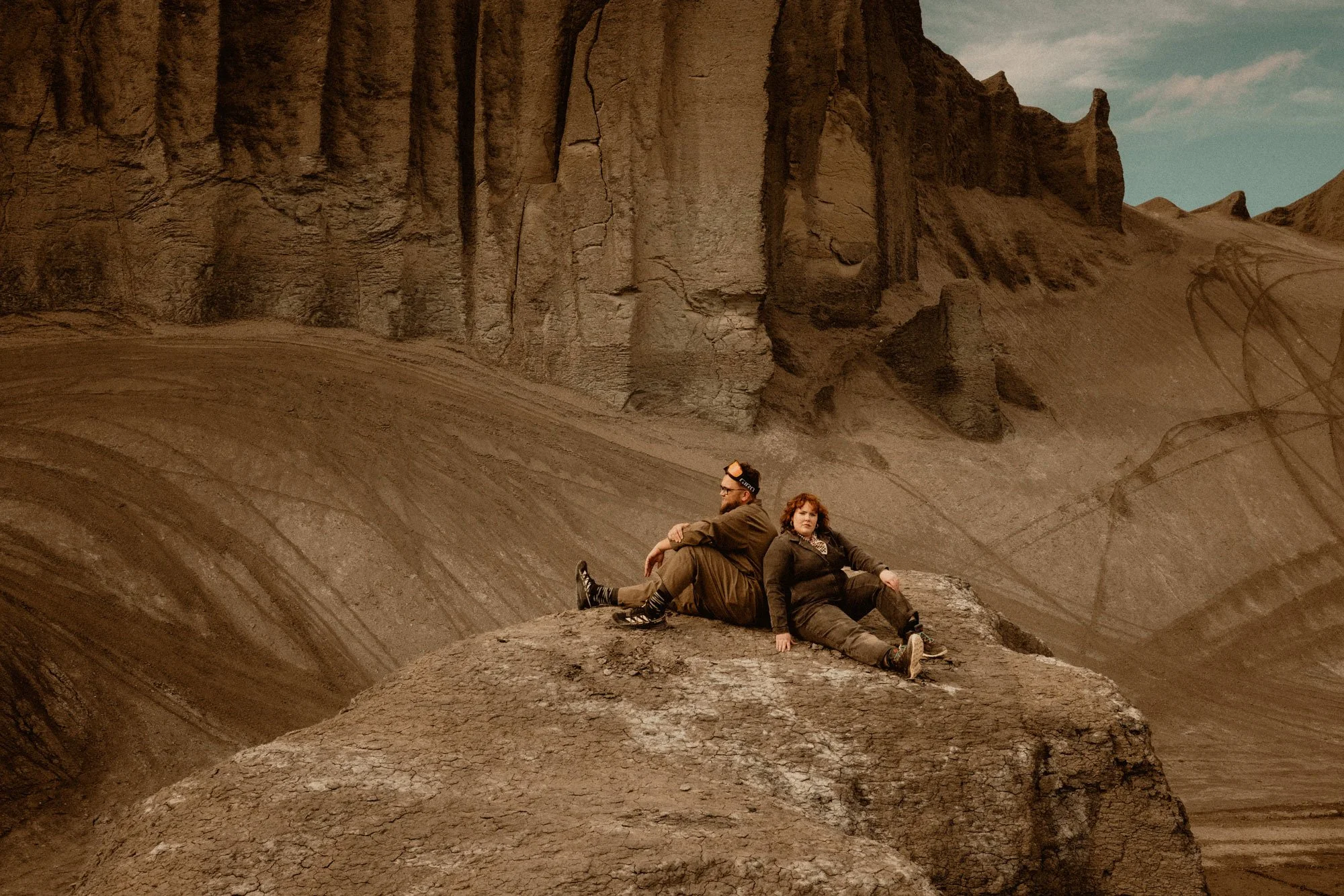 Two people sitting on a large rock in the Utah desert with steep, layered cliffs in the background.