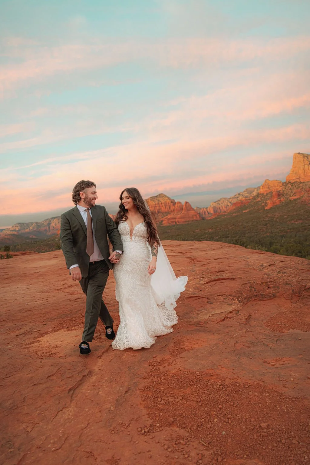 A bride and groom holding hands and walking on red rock terrain at sunset with distant red rock formations and a colorful sky.