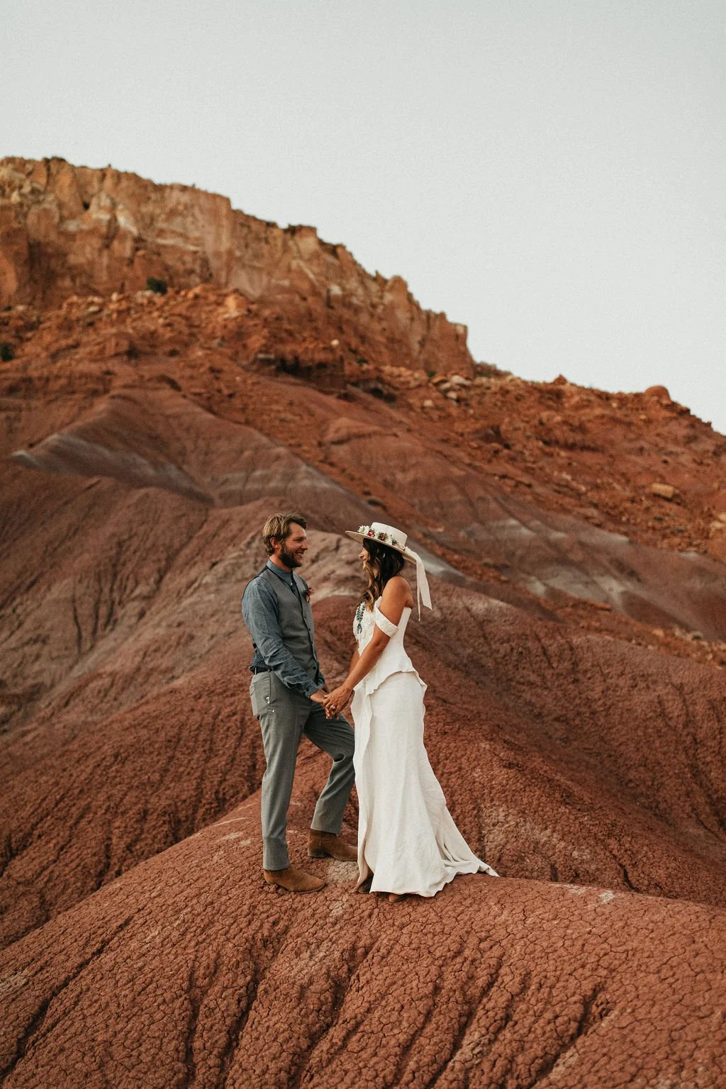 A couple dressed in wedding attire standing on a reddish-brown rocky landscape with a mountain in the background, holding hands and smiling at each other.