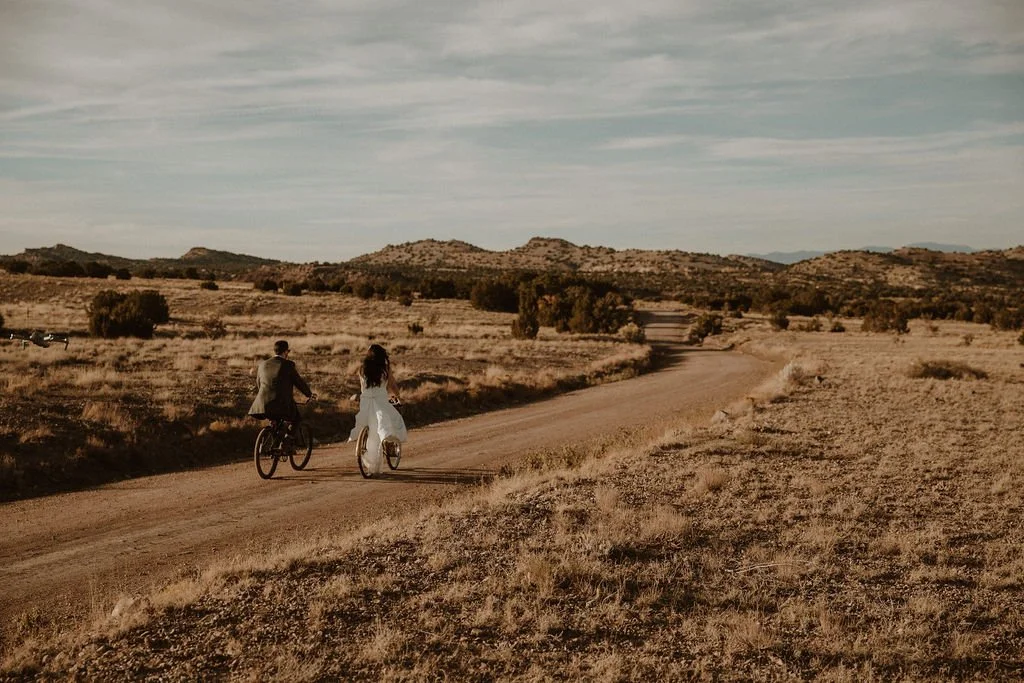 A man and woman riding bicycles on a dirt trail in a dry, open landscape with hills and sparse trees under a cloudy sky.