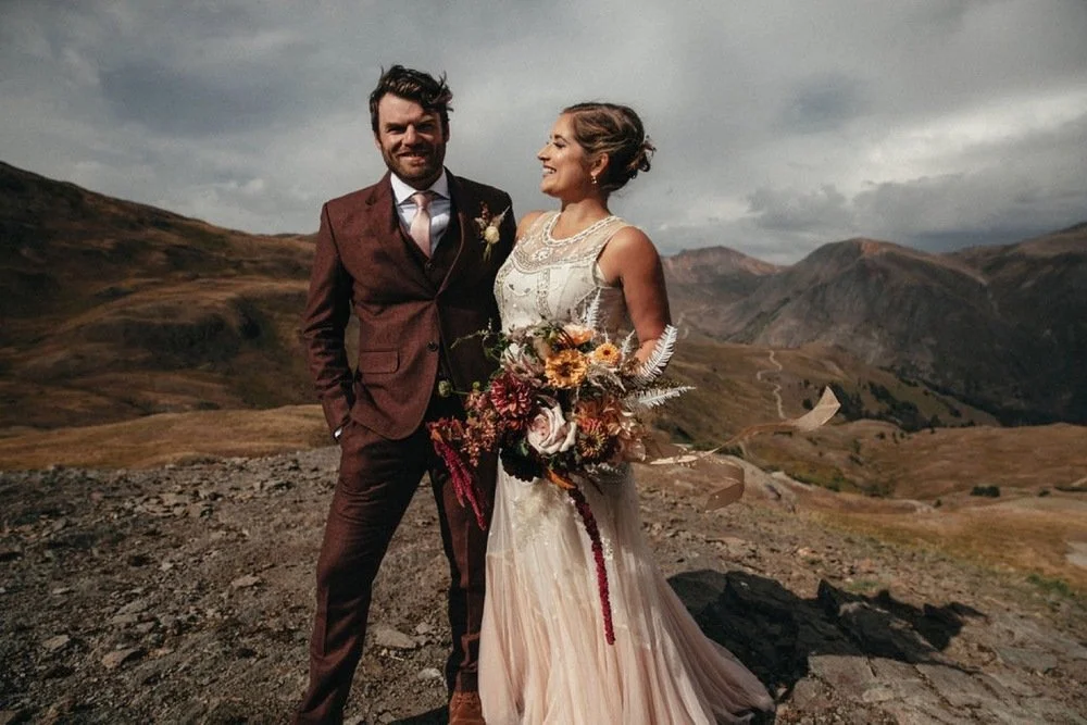 A bride and groom stand together outdoors on rocky terrain with mountainous landscape in the background, dressed in wedding attire, smiling and looking at each other.