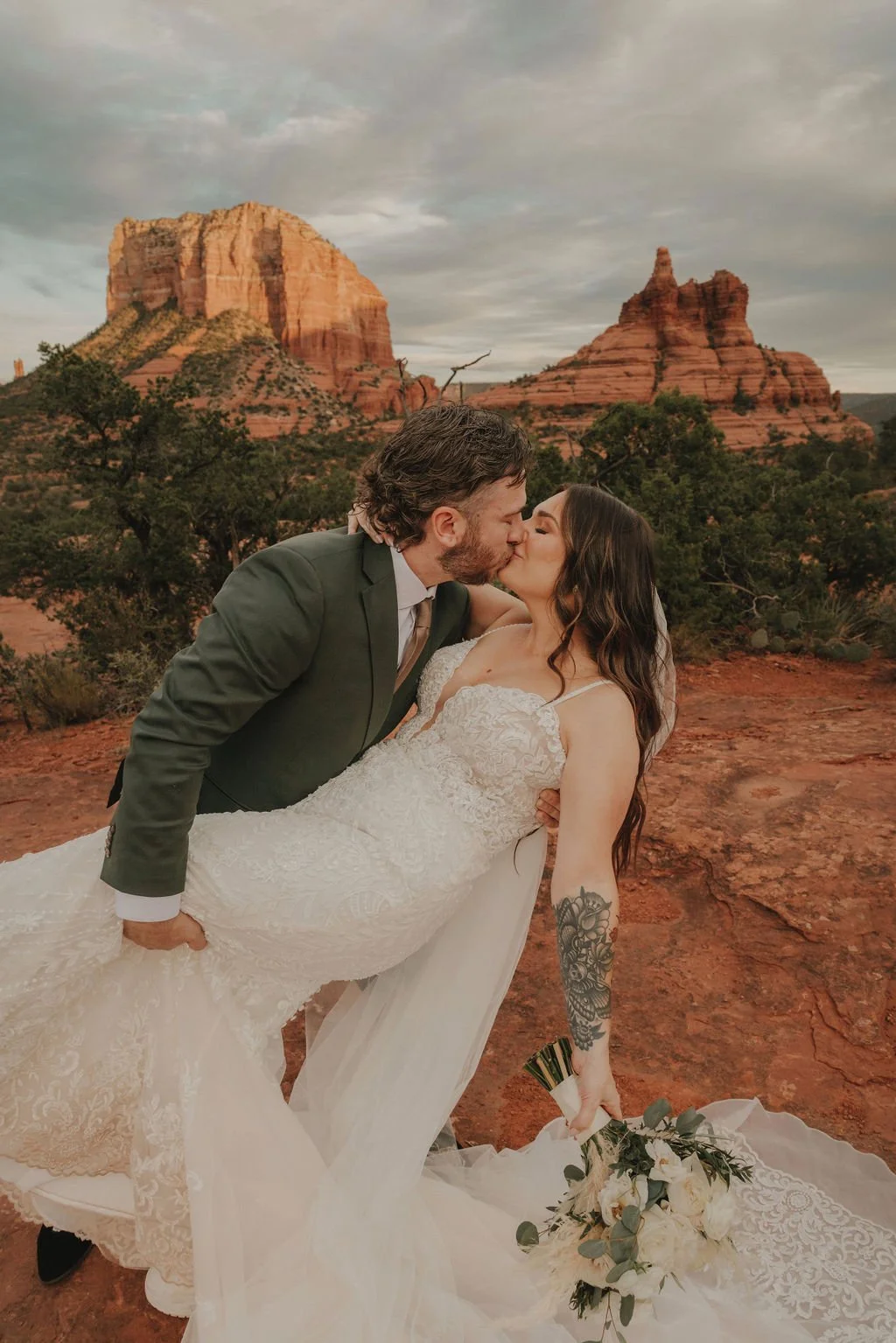 A bride and groom kissing in front of red rock formations at sunset, with the bride holding a bouquet of white roses and greenery.
