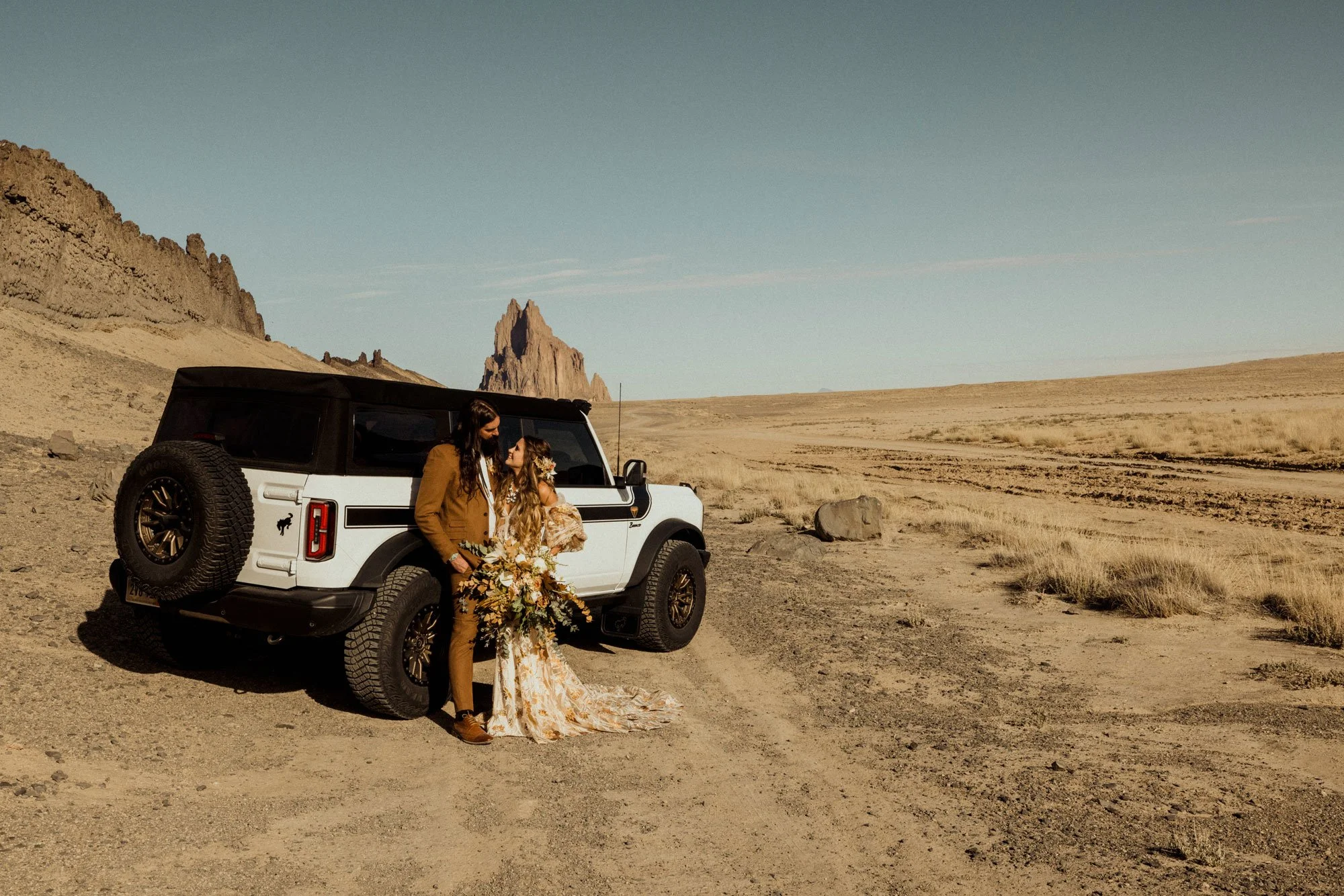 A couple dressed in wedding attire standing beside a white SUV in a desert landscape with distant rocky formations and clear blue sky.