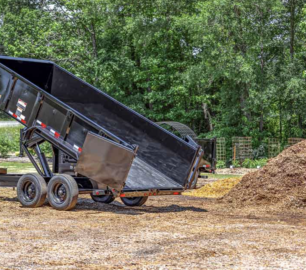 A black dump trailer with a raised bed on a gravel surface, surrounded by green trees and a mound of dirt nearby.