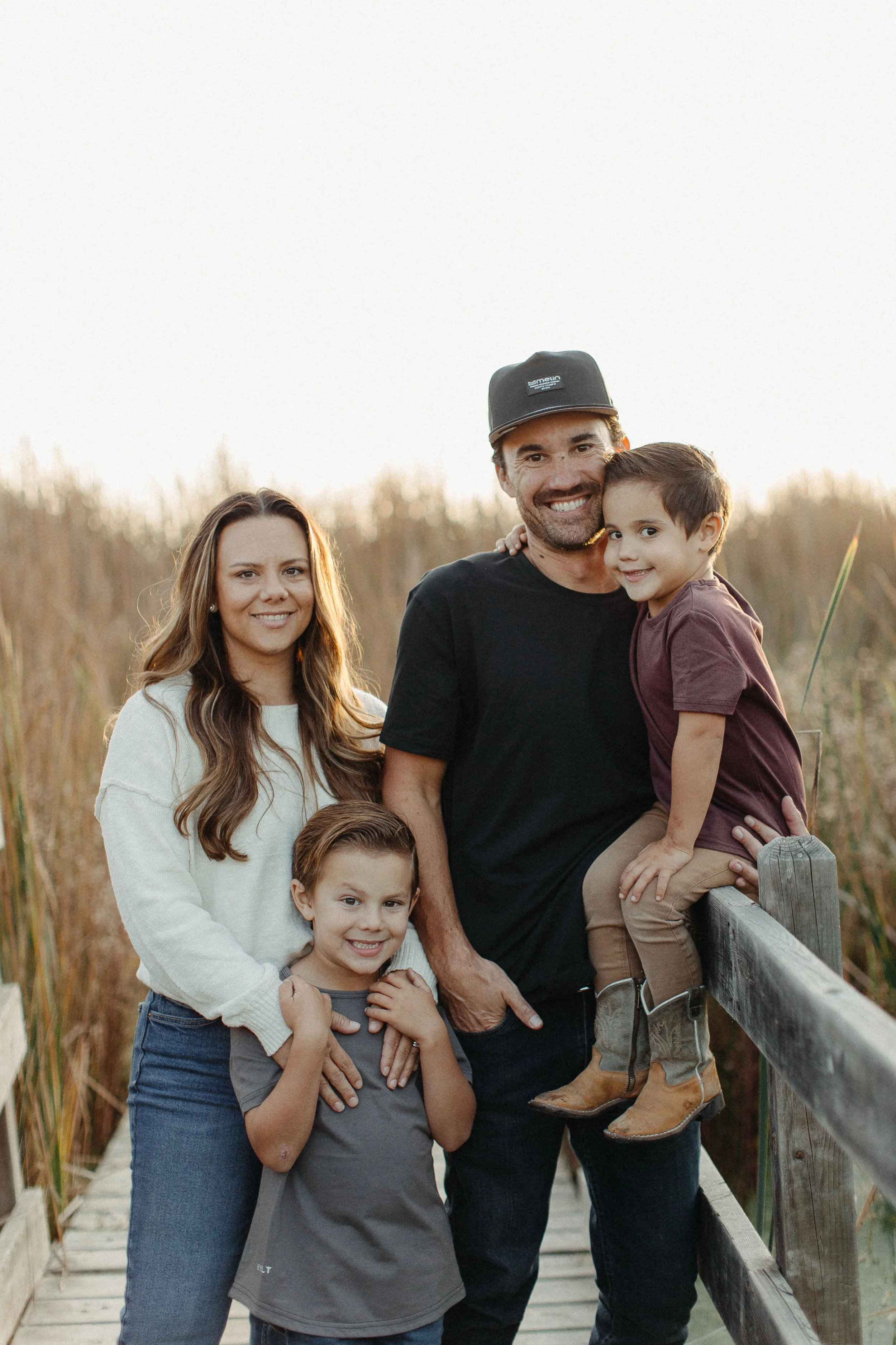 A family of five standing on a wooden bridge outdoors at sunset, all smiling and looking at the camera.
