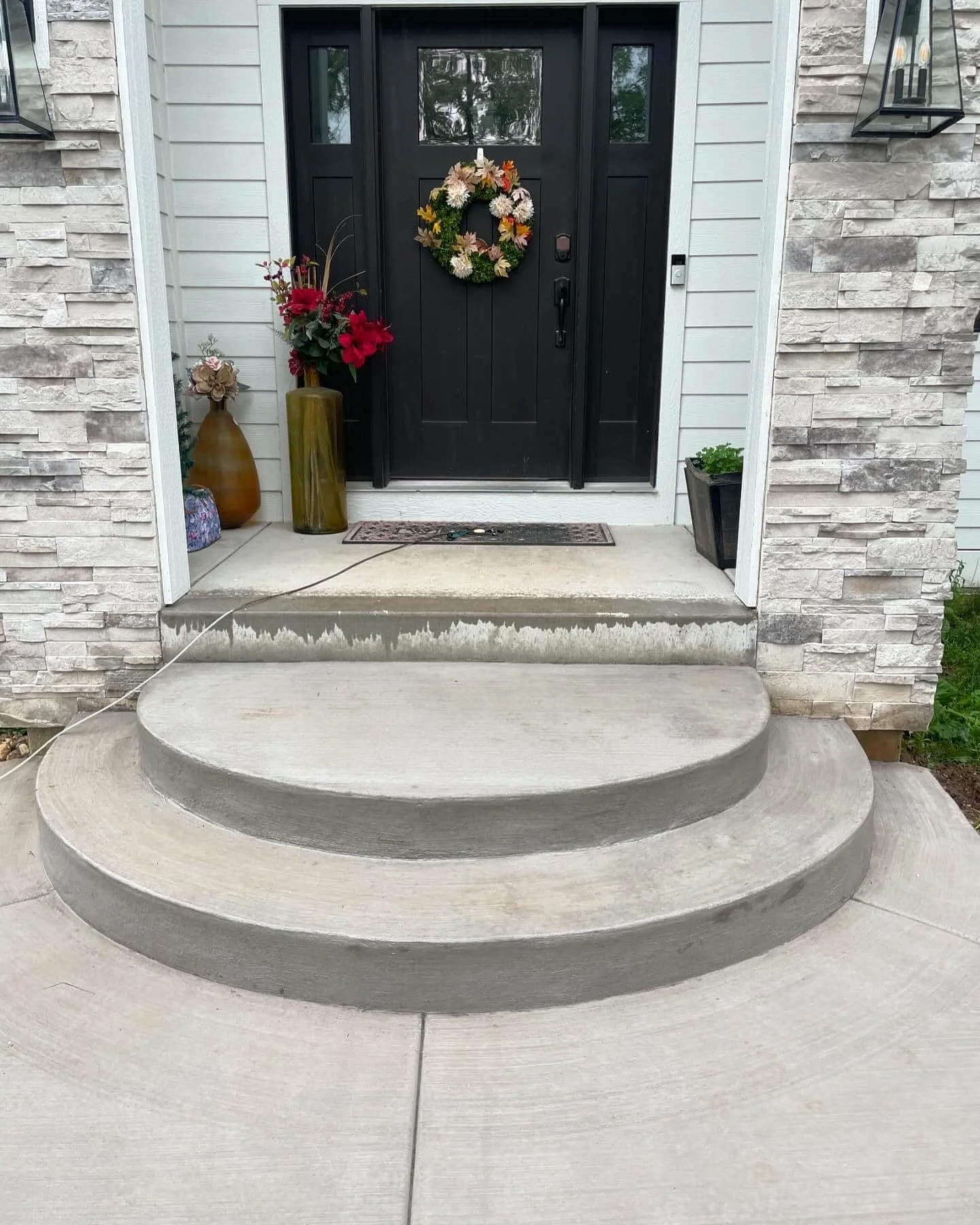 Concrete steps leading up to a black front door decorated with a flower wreath, surrounded by potted plants and vases.